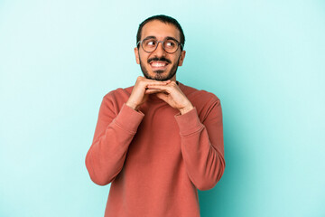 Young caucasian man isolated on blue background keeps hands under chin, is looking happily aside.