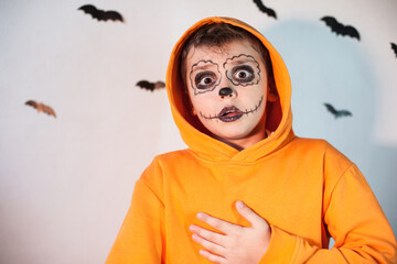 a boy in an orange sweatshirt with Halloween makeup stands with a surprised face against the background of a wall with bats