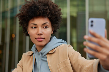 Adorable young Afro American woman clicks sefie images during free time in city uses smartphone...