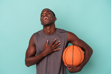 Young African American man playing basketball isolated on blue background laughs out loudly keeping hand on chest.