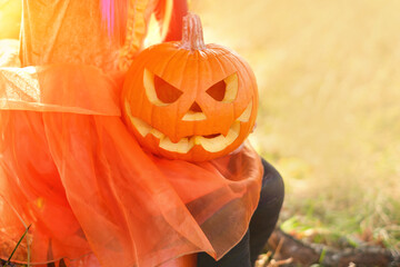 Pumpkin with an evil face in the hands of a child in the rays of the setting sun on Halloween. Traditions of carving pumpkins and asking for treats and sweets on Halloween