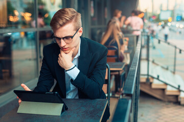 Young handsome blond guy sits with a tablet on the terrace of a cafe, works or studies in a public place