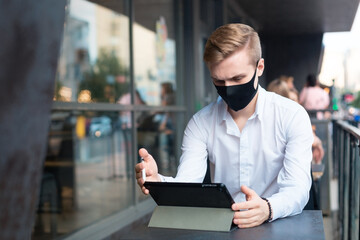 Young handsome blond guy sitting in a mask and working during a pandemic in a public place