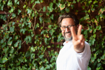 Happy mature man wearing eyeglasses and showing victory fingers sign while standing against...