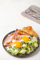 Continental breakfast - eggs, bacon, micro greens and cucumber slices on a plate on the table. Healthy breakfast.
