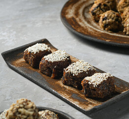 A sweet dessert of dried fruits covered with sesame seeds lies on a ceramic stand, next to oatmeal balls in a plate. Close-up, healthy eating, trend, selective focus..