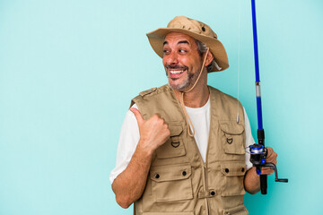 Fototapeta premium Middle age caucasian fisherman holding rod isolated on blue background points with thumb finger away, laughing and carefree.