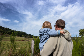 Little girl sitting on her father's arms and enjoying rural view of the green field with cows.