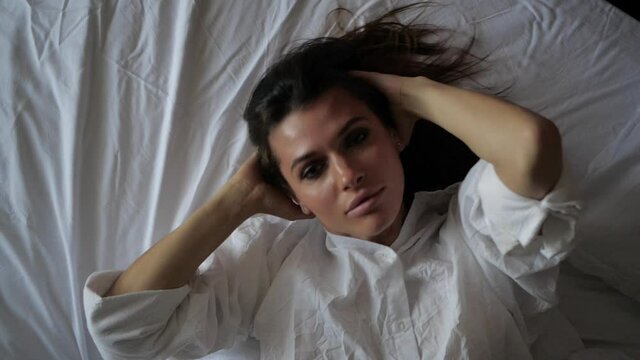 sexy brunette girl lies on a white bed, plays with her hair and looks straight into the camera. High quality FullHD footage