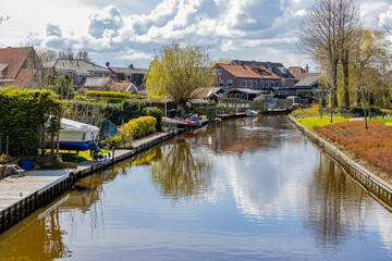 Canal between gardens, bare trees, green grass, boats and houses in the background, sunny spring day with a blue sky and white clouds at Medemblik in Noord-Holland, Netherlands