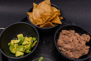 Bowls of tortilla chips, chopped avocado, and refried beans on Mexican comal