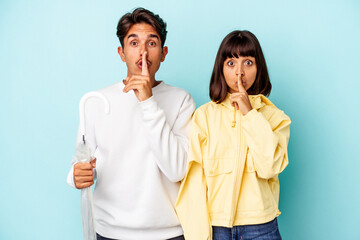 Young mixed race couple holding umbrella isolated on blue background keeping a secret or asking for silence.
