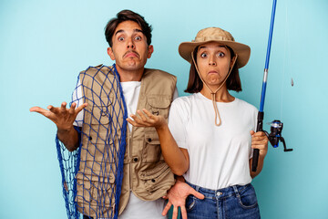 Young mixed race fisher couple isolated on blue background shrugs shoulders and open eyes confused.