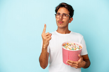 Young mixed race man eating popcorns isolated on blue background showing number one with finger.