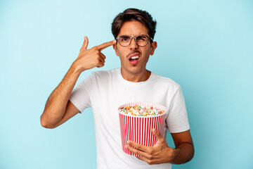 Young mixed race man eating popcorns isolated on blue background showing a disappointment gesture with forefinger.