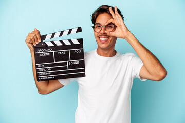 Young mixed race man holding clapperboard isolated on blue background excited keeping ok gesture on...