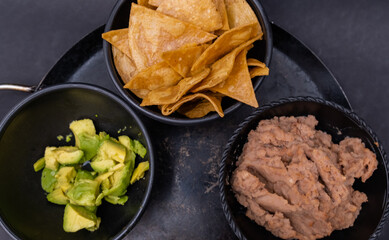 Bowls of tortilla chips, chopped avocado, and refried beans on Mexican comal
