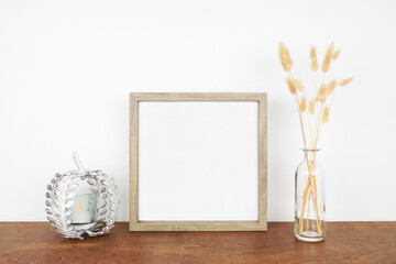 Mock up square wooden frame with fall grasses and pumpkin candle holder on a wood shelf against a...