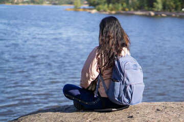 Young brunette woman sits with her back to the camera, backpack behind her shoulders and looks at the panorama of the lake. Concept of relaxation in nature.