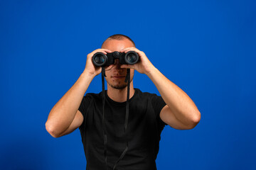 Young man over isolated blue wall and looking in the distance with binoculars.