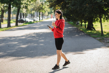 Stylish girl in red sweater holding smartphone and drinking coffee. Happy woman with beautiful smile chatting on cell smartphone while relaxing in the park, outdoors