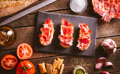 Iberian ham toasts on a slate plate on a brown wooden table with some ingredientes around it....