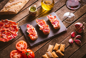 Iberian ham toasts on a slate plate on a brown wooden table with some ingredientes around it....