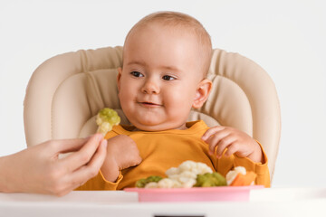 Mom's hand gives a baby sitting on a feeding chair boiled broccoli
