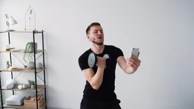 Vain sport. Posing man. Fitness blogging. home training. Funny selfish athletic guy making selfie with dumbbells in hand on mobile phone playing fancy in light room interior.