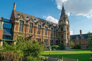 View of historical bricked Christ's College building attached to a medieval style clock tower in front of lawn and benches in Cambridge England