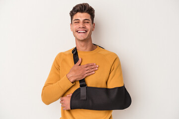 Young caucasian man with broken hand isolated on white background laughs out loudly keeping hand on chest.