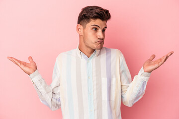 Young caucasian man isolated on pink background confused and doubtful shrugging shoulders to hold a...