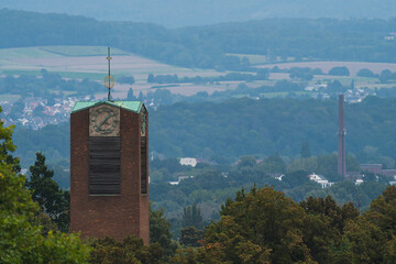 a church tower between trees and the city, hills and forest in the background 