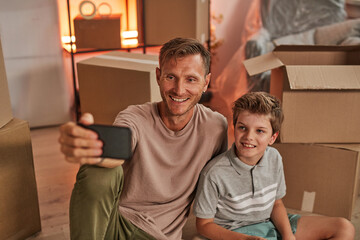 Fototapeta na wymiar Portrait of father and son taking selfie photo with cardboard boxes while moving in to new home, copy space