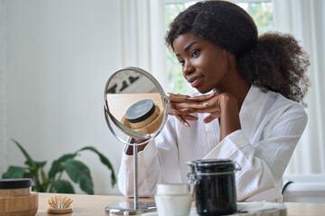 Happy pretty smiling young black woman model doing makeup or skin care morning routine looking in...