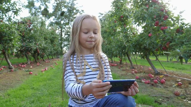 Little cute girl on nature apple tree garden sit watch smartphone while have video chat