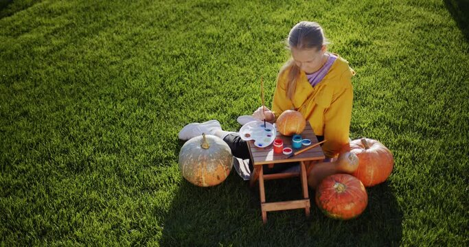 Top view: A child paints a pumpkin, prepares decorations for Halloween. Sits on the lawn in the backyard of the house