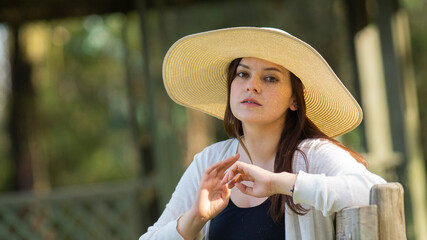 Portrait of beautiful long-haired Hispanic young woman wearing a hat sitting on a park bench with a pensive attitude against a background of unfocused green trees