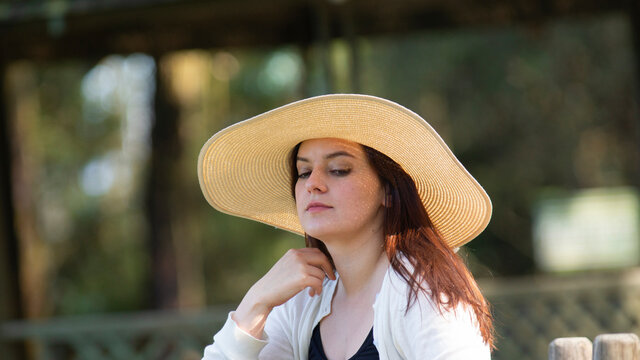 Portrait of beautiful long-haired Hispanic young woman wearing a hat sitting on a park bench with a pensive attitude against a background of unfocused green trees - Powered by Adobe