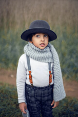 boy child in plaid pants, hat, suspenders and scarf stands in a field