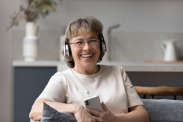 Happy laughing middle aged old woman in eyewear listening f music or affirmations in headphones,...