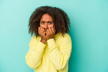 Young african american woman with curly hair isolated on blue background biting fingernails,...