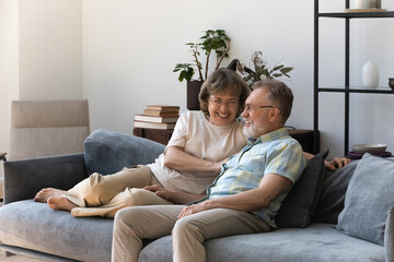 Affectionate smiling middle aged couple enjoying talking communicating resting on comfortable sofa in modern living room, sincere loving bonding relations of retired couple, ageing together concept.