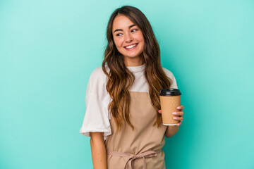 Young caucasian store clerk woman holding a takeaway coffee isolated on blue background looks aside smiling, cheerful and pleasant.