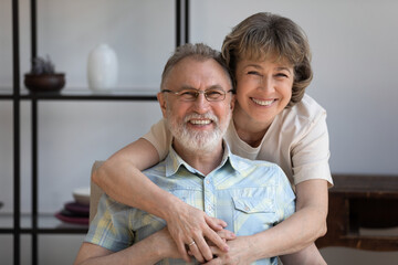 Portrait of affectionate mature woman cuddling shoulders of smiling senior husband, resting on cozy...