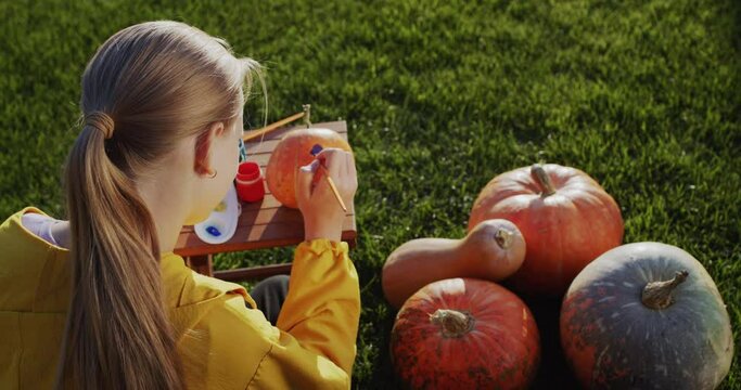 Rear view: A child paints a pumpkin, prepares decorations for Halloween. Sits on the lawn in the backyard of the house