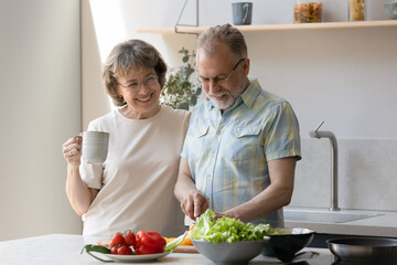Smiling beautiful old mature woman drinking hot coffee or tea, standing near loving middle aged husband chopping fresh vegetables for salad, preparing healthy breakfast on weekend i modern kitchen.