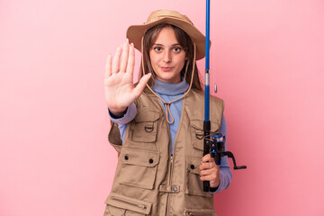 Young caucasian fisherwoman holding a rod isolated on pink background standing with outstretched hand showing stop sign, preventing you.