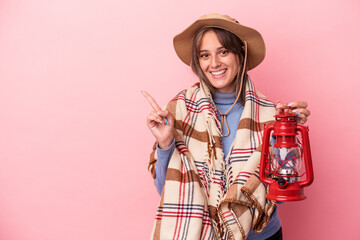 Young caucasian woman holding vintage lantern isolated on pink background smiling and pointing aside, showing something at blank space.