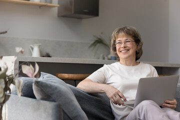 Dreamy senior middle aged woman in glasses looking in distance, holding computer on laps, planning vacation time, enjoying communicating in social networks, older people and modern technology concept.